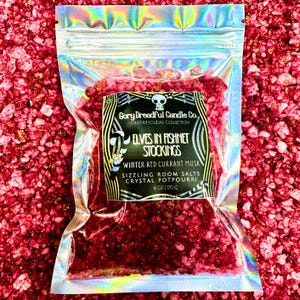 Op de afbeelding: Een doorzichtige, iriserende zak gevuld met rode kristallen potpourri. Het etiket luidt "Elves in Fishnet Stockings" en "Winter Red Currant Musk". Het product is van Gory Dreadful Candle Co. en weegt 170 g.