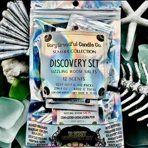 Puede incluir: Un paquete transparente e iridiscente que contiene un "Discovery Set" de sales de baño efervescentes de Gory Dreadful Candle Co. El set incluye 12 aromas, con un peso total de 238,2 gramos. El embalaje presenta texto, incluyendo nombres de aromas y la frase "Summer Collection."