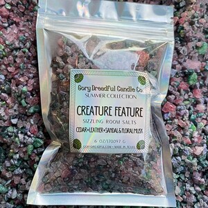 Pode incluir: Um pequeno saco de plástico transparente cheio de sais de banho coloridos. O rótulo diz "Gory Dreadful Candle Co. Summer Collection Creature Feature Sizzling Room Salts Cedar+Leather+Sandal & Floral Musk 6 oz/170097 g GoryDreadful.com Made in Texas"