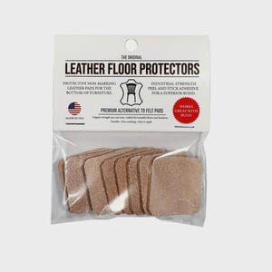 Op de afbeelding: Een verpakking met lederen vloerbeschermers. De verpakking toont de tekst "LEATHER FLOOR PROTECTORS". De verpakking bevat meerdere vierkante, lichtbruine leren pads. De pads zijn een premium alternatief voor vilt pads. Er staat "MADE IN USA" op.