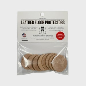 Può includere: Una confezione di protezioni per pavimenti in pelle. La confezione mostra il testo "Leather Floor Protectors" e "Made in USA". La confezione contiene diversi cuscinetti rotondi in pelle marrone chiaro. I cuscinetti sono un'alternativa premium ai feltrini.
