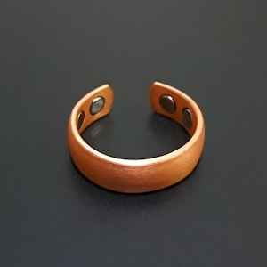 Könnte beinhalten: Ein kupferfarbener, verstellbarer Ring mit drei kleinen, runden Magneten. Der Ring hat eine glatte, matte Oberfläche und ein schlichtes, offenes Design. Der Ring wird vor einem dunklen Hintergrund gezeigt.