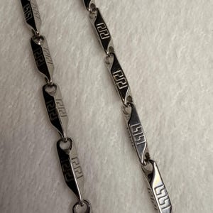 May include: A silver-toned metal chain necklace featuring a series of elongated, rectangular links. Each link is polished and engraved with a repeating geometric Greek key pattern in black, connected by small circular jump rings in a linear design.