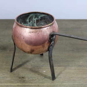 Op de afbeelding: Een kleine koperen pot met een handvat en drie ijzeren poten. De pot heeft een versleten, verweerde uitstraling.