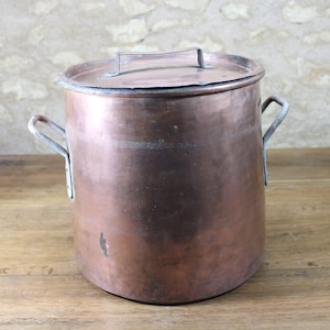 Op de afbeelding: Een grote koperen pot met twee handvatten en een deksel. De pot staat op een houten oppervlak.