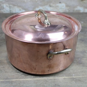 Op de afbeelding: Een koperen pan met een deksel en twee messing handvatten. De pan staat op een houten oppervlak.
