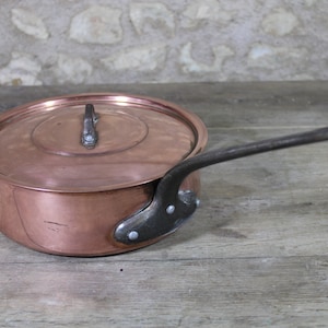 Può includere: Una pentola di rame con un coperchio e un manico in ferro nero. La pentola ha una finitura martellata ed è appoggiata su una superficie di legno.