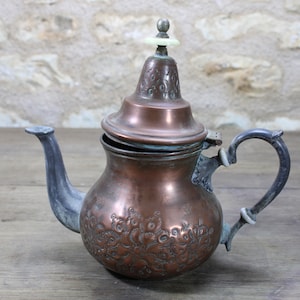 May include: A copper-colored teapot with a decorative floral pattern on the body. The teapot has a curved spout and a handle with a loop design. The lid is topped with a small, light-colored finial. The teapot is likely vintage or antique.
