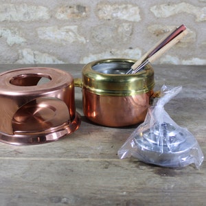 Op de afbeelding: Een koperen fonduepan met een messing rand en een houten handvat. De pan staat op een koperen standaard met een gat in het midden. Er zitten drie houten fonduevorken in de pan. De pan staat op een houten oppervlak.