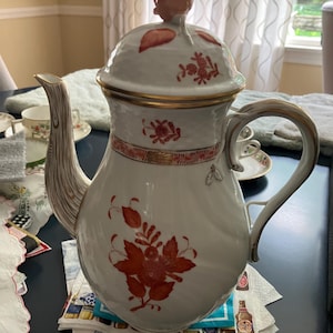 May include: A white porcelain teapot with a lid and handle, adorned with red floral patterns and gold trim. The spout has a textured, branch-like design, and the lid features a rose-shaped finial. A decorative piece for serving tea.