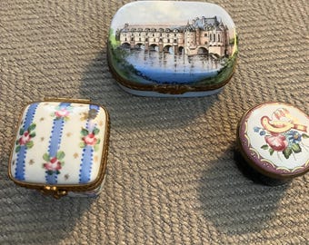May include: Three decorative trinket boxes. One rectangular box features a painting of a castle over water. Another square box has a floral and striped design. A round box with a floral design is also present.