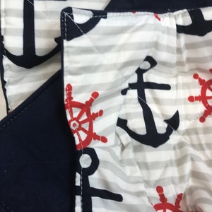 May include: Close-up of a white and gray striped fabric with a navy blue anchor and red steering wheel pattern. The fabric is quilted and has a navy blue border.
