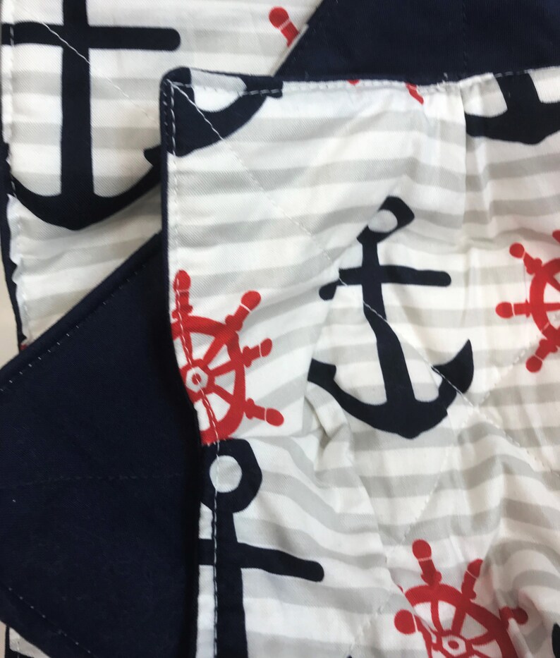 May include: Close-up of a white and gray striped fabric with a navy blue anchor and red steering wheel pattern. The fabric is quilted and has a navy blue border.