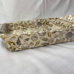May include: A beige fabric baking dish with a glass insert and a seashell pattern. The dish has two handles on the sides.