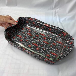 May include: A black and white fabric-covered baking dish with a clear glass insert. The fabric has a repeating pattern of red trucks with the text "Jingle Bells All the Way" and "Dashing Through the Snow".