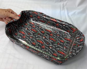 May include: A black and white fabric-covered baking dish with a clear glass insert. The fabric has a repeating pattern of red trucks with the text "Jingle Bells All the Way" and "Dashing Through the Snow".