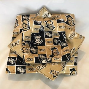 May include: A set of three black and gold fabric pot holders with a repeating pattern of the University of Central Florida (UCF) logo and knight mascot. The pot holders are folded into a star shape.