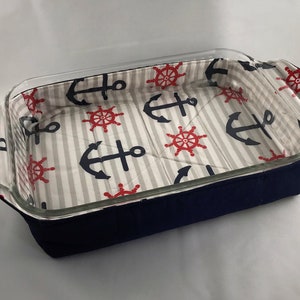 May include: A navy blue fabric baking dish liner with a white and gray striped background. The liner features a nautical pattern of red ship wheels and blue anchors.