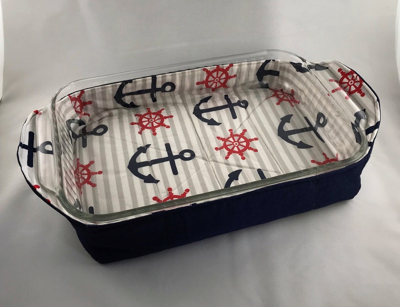 May include: A navy blue fabric baking dish liner with a white and gray striped background. The liner features a nautical pattern of red ship wheels and blue anchors.