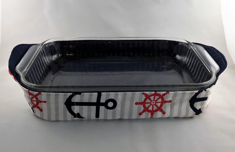 May include: A glass baking dish with a navy blue and white striped fabric cover. The fabric has a nautical theme with anchors and ship wheels.