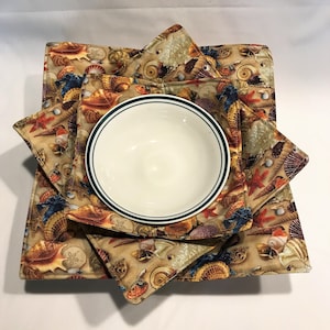 May include: Set of three square fabric bowl cozies with a seashell pattern. The cozies are folded and stacked with a white bowl inside.