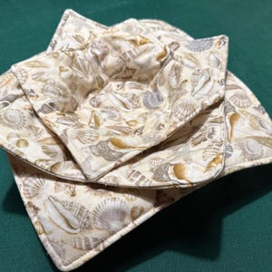 Puede incluir: Una pila de tres salvamanteles cuadrados de tela. Los salvamanteles son de color beige con un estampado de conchas marinas en tonos marrones, beiges y grises. Los bordes están acabados con un ribete blanco. Están diseñados para sujetar cuencos.