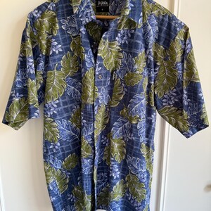 May include: Blue button-down shirt with a green and white leaf pattern. The short-sleeved shirt has a collared neckline and a chest pocket. The shirt is made of a lightweight fabric and is hanging on a wooden hanger.
