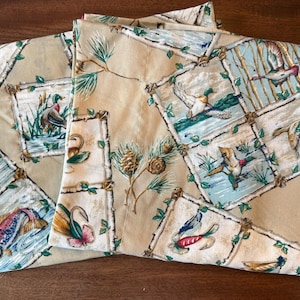 May include: Tan fabric with a repeating pattern of framed nature scenes. Each frame features a different image of fish, ducks, and other wildlife. Green pine needles and pine cones are also featured in the design.