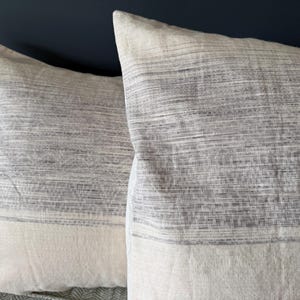 May include: Two square decorative pillows. The pillows have a light gray and white striped pattern on the top half and a solid white bottom half. The pillows are made of a soft, textured fabric.