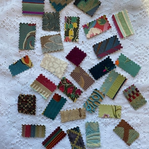 May include: A collection of fabric swatches in various colours and patterns, including teal, green, brown, red, and purple. The swatches are arranged on a white lace tablecloth.