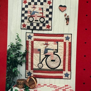 May include: Two patriotic quilts with a bicycle and tractor design, featuring red, white, and blue colors. The quilts are square-shaped, with star and checkerboard patterns. A heart-shaped decoration and a small figurine are also visible.