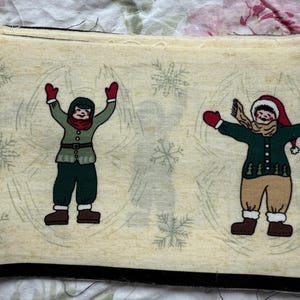 May include: Cream-colored fabric with a winter theme. The fabric features illustrations of children in winter attire, including red mittens and hats, green coats, and brown boots. Snowflakes are scattered across the background.