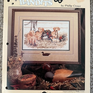 May include: A framed print titled "The Little Bandits" by Phillip Crowe, featuring a cross-stitch design of several golden retriever puppies and ducks. The image is surrounded by a decorative border and includes a decorative duck decoy and a tin.