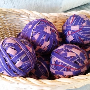 May include: A wicker basket filled with purple and pink yarn balls. The yarn is made of fabric strips and is tightly wound into balls.