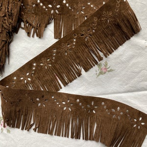 May include: Brown suede fringe trim with laser-cut floral and dot designs. The trim is in a roll and laid out flat, showing the decorative cutouts and the fringe detail. This trim is suitable for various craft and sewing projects.