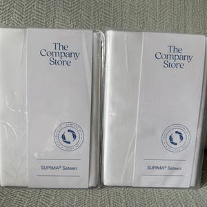 May include: Two white, rectangular packages wrapped in clear plastic. Each package has a white label with the text "The Company Store" in blue, along with a circular logo and the words "SUPIMA Sateen".