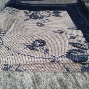 May include: A folded beige throw blanket with a navy blue border and a floral pattern. The blanket is made of a soft, textured fabric.
