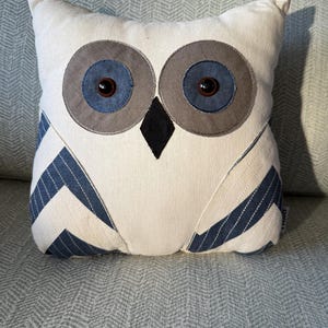 May include: A decorative owl-shaped pillow with a white body, gray and blue eyes, and a black beak. The wings feature blue and white striped fabric. The pillow is designed to resemble an owl.