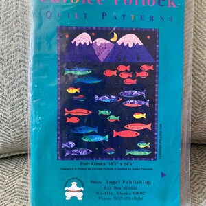 May include: A teal-colored package featuring a quilt pattern titled "Fish Alaska." The quilt design includes colorful fish, mountains, and a moon. The pattern measures 18.5" x 24.5" and is designed by Carolee Pollock.