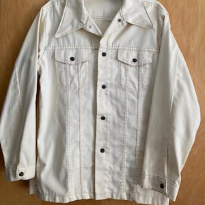 May include: A white denim jacket with a button-up front and two chest pockets. The jacket has brown stitching and black buttons.