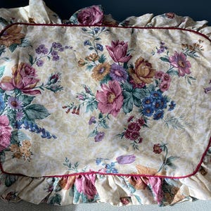 May include: A cream-colored pillowcase with a floral print featuring pink, red, blue, and yellow flowers. The pillowcase has a ruffled edge and a burgundy trim.