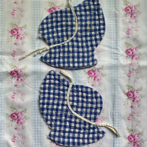 May include: Two blue and white gingham fabric bonnet caps with white ties. The caps are quilted and have a scalloped edge.