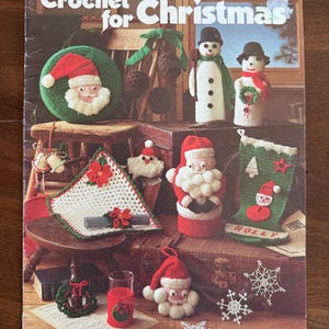 May include: A vintage book cover titled "Crochet for Christmas" featuring various crocheted Christmas decorations. The cover displays a Santa Claus face, snowmen, a stocking, and other festive items in red, green, and white.