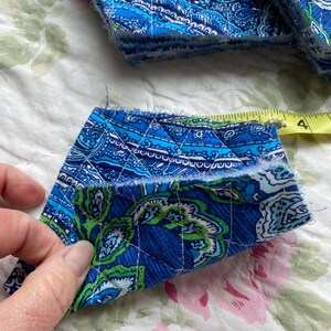 May include: A stack of quilted fabric squares with a blue paisley pattern and white stitching. One square is held up, showing the pattern and a measurement of approximately 3 inches. The fabric has green and white accents.