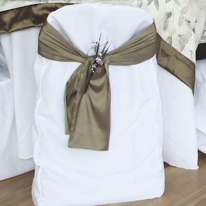 May include: White chair cover with a brown satin sash tied in a bow. A small floral accent is pinned to the bow. The chair is set for a wedding or special event. The table cloth has a floral pattern.