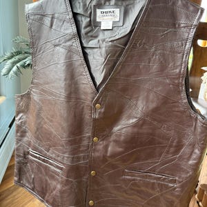 May include: A dark brown leather vest featuring a patchwork design of stitched leather panels. The vest has a V-neckline, four brass-colored metal snap buttons down the front, and two side pockets. A label inside reads DUKE HABAND.