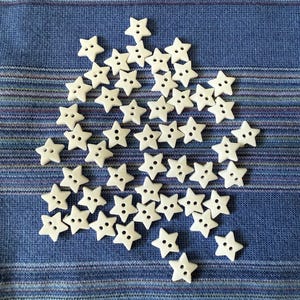 May include: A collection of small, white star-shaped buttons scattered on a blue striped fabric. Each button has two holes for sewing. The buttons are a craft supply, suitable for various sewing projects.