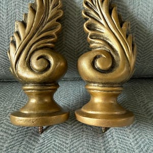 May include: A pair of gold-coloured, decorative finials featuring a leaf design. Each finial has a rounded base and a screw for attachment. The finials are displayed on a blue-grey patterned fabric.