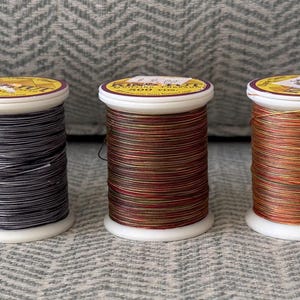 May include: A row of seven thread spools in a range of colours: green, purple, blue, and browns. Each spool has a white base and top, with a label. The threads are tightly wound, ideal for sewing or craft projects.