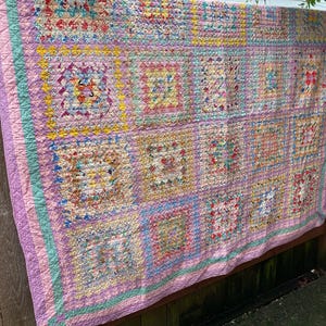 May include: A pink and green patchwork quilt with a variety of colorful squares and patterns. The quilt is hanging on a wooden fence.